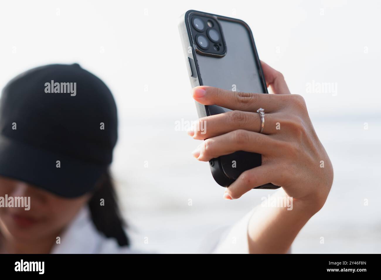 Gros plan d'une main tenant un téléphone intelligent, avec un focus sur une bague sur le doigt. L'arrière-plan présente une scène de plage floue, suggérant un o décontracté Banque D'Images