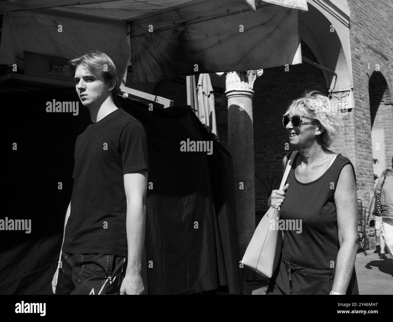 Cremona, Italie 31 août 2024 Un jeune homme et une femme mature marchant dans une rue de la ville devant des étals de marché, baignés de soleil brillant Banque D'Images