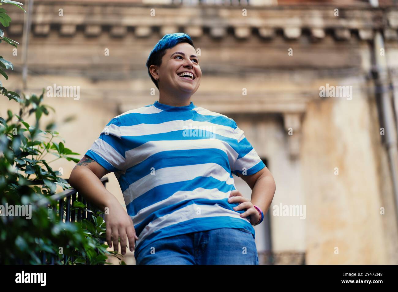 Individu joyeux de genre non conforme avec les cheveux bleus courts et chemise rayée riant à l'extérieur. Expression authentique du bonheur et de la confiance en soi Banque D'Images