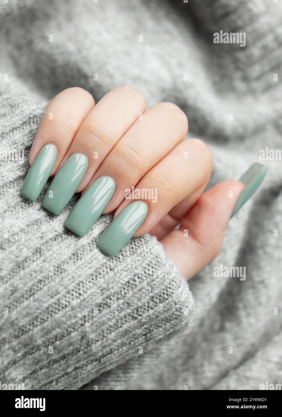 Une main élégante avec des ongles vert menthe bien manucurés est délicatement placée sur un pull gris doux, mettant en valeur un accent à la mode dans un environnement confortable. Banque D'Images