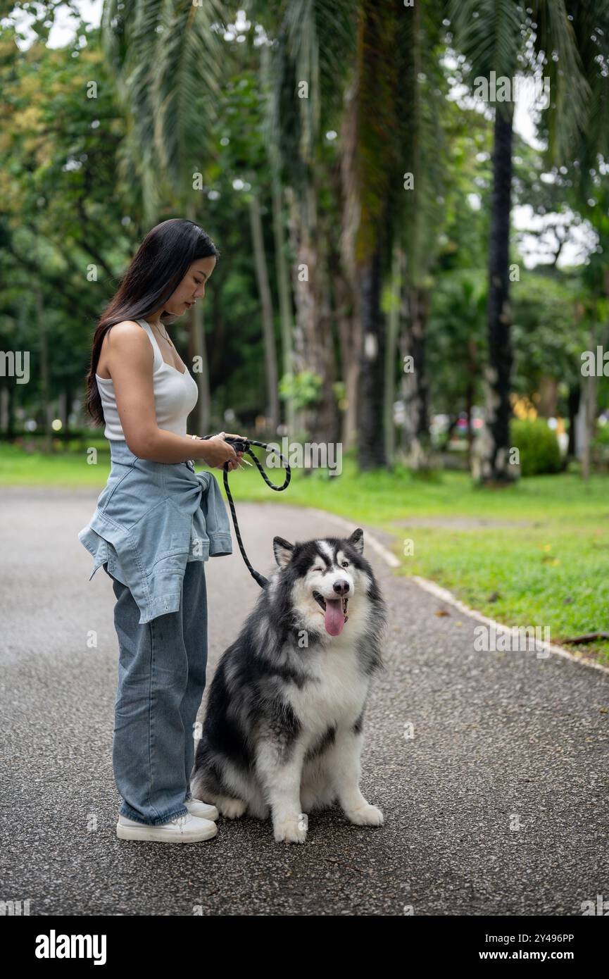 Une belle femme asiatique emmène son chien se promener en laisse dans un parc verdoyant, l'entraînant à s'asseoir et à obéir tout en jouant à l'extérieur. chien et propriétaire con Banque D'Images