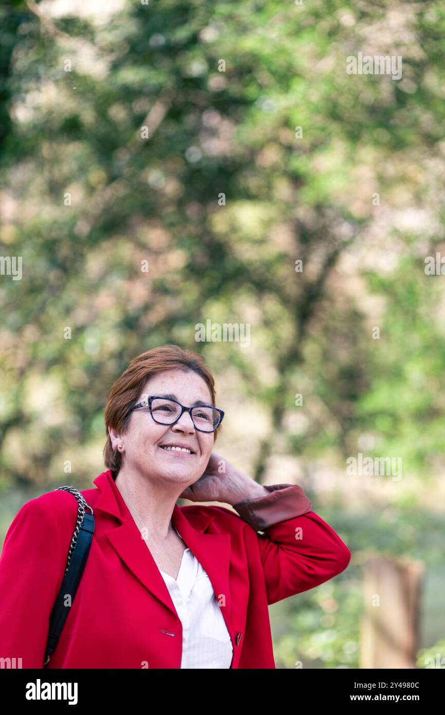 Une femme mature avec des lunettes sourit chaudement tout en portant un blazer rouge. L'arrière-plan est flou, avec un feuillage vert luxuriant et la lumière du soleil filtrant à travers Banque D'Images