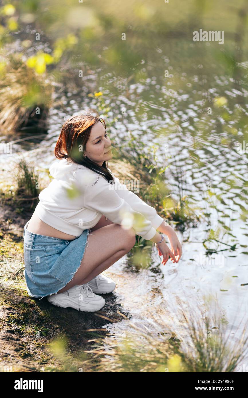 Portrait d'une jeune femme aux cheveux roux à travers les feuilles des arbres. Elle squat près d'un bord de lac tranquille pour enjouer la nature. Banque D'Images