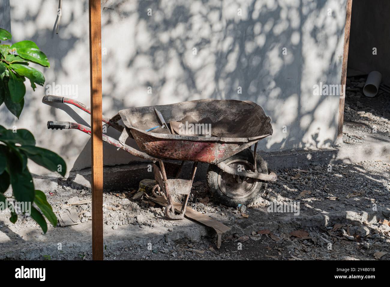 Une brouette rouge altérée repose sur une surface de gravier, partiellement ombragée par des arbres. La brouette est remplie de saleté et d'outils, avec un mur dans le bac Banque D'Images
