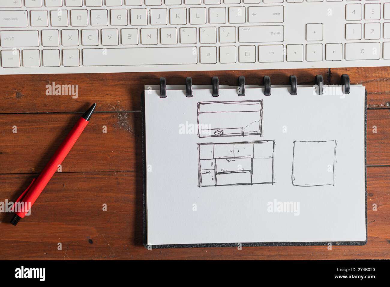 Un carnet de croquis avec des dessins dessinés à la main, y compris une mise en page avec une télévision et des meubles, placé sur une table en bois à côté d'un stylo rouge et d'un clavier d'ordinateur. Banque D'Images