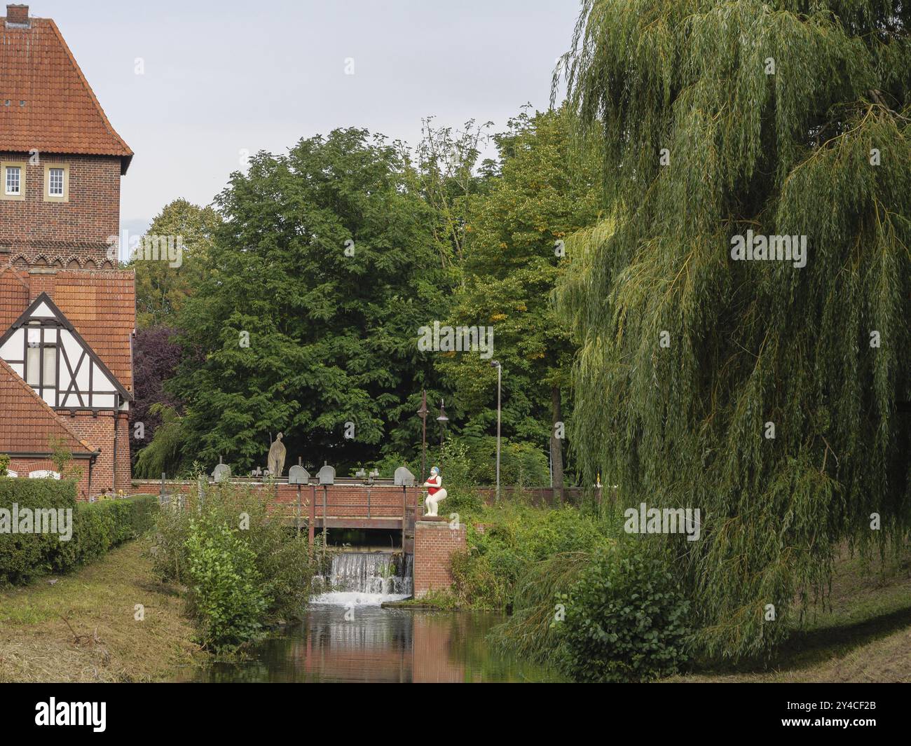 Cours de rivière avec saule et sculpture devant un bâtiment historique en briques dans un cadre verdoyant, Coesfeld, muensterland, allemagne Banque D'Images