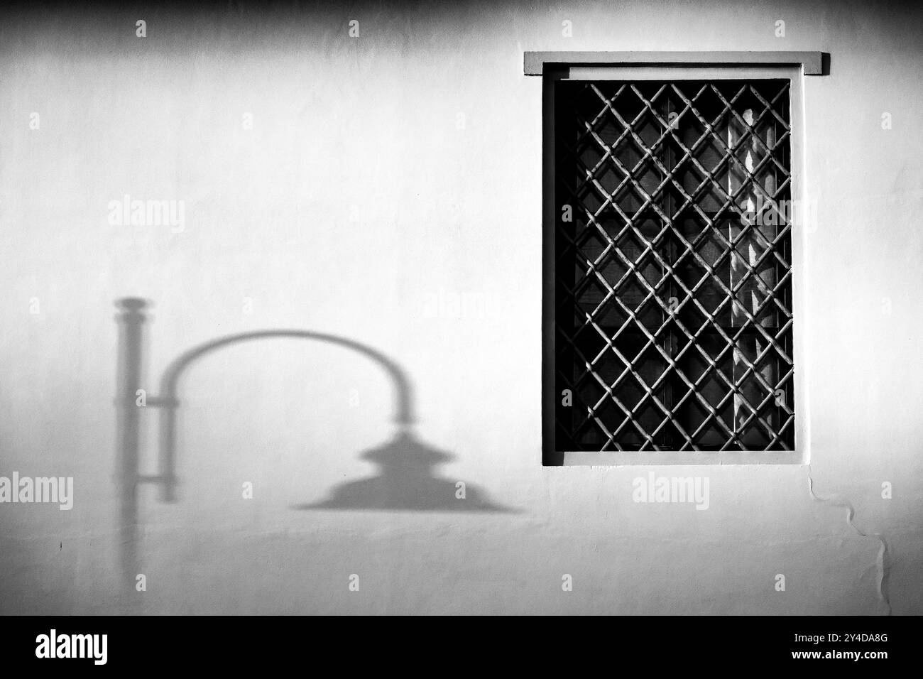 Photographie en noir et blanc d'une fenêtre avec des barres de treillis et l'ombre d'un lampadaire vintage sur un mur blanc. Banque D'Images