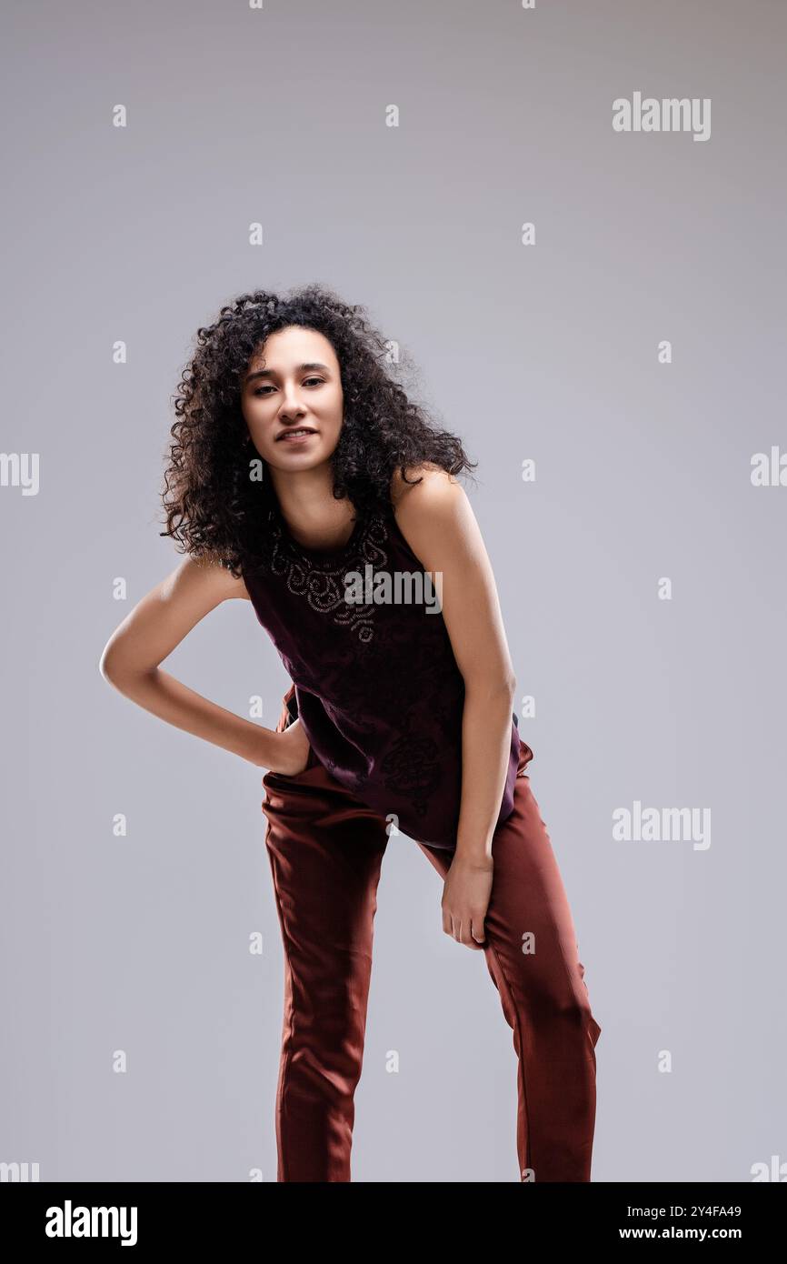 Une jeune femme aux cheveux bouclés pose avec une main sur sa hanche et l'autre sur son genou. Créer une pose dynamique et confiante. Sa tête est inclinée sl Banque D'Images