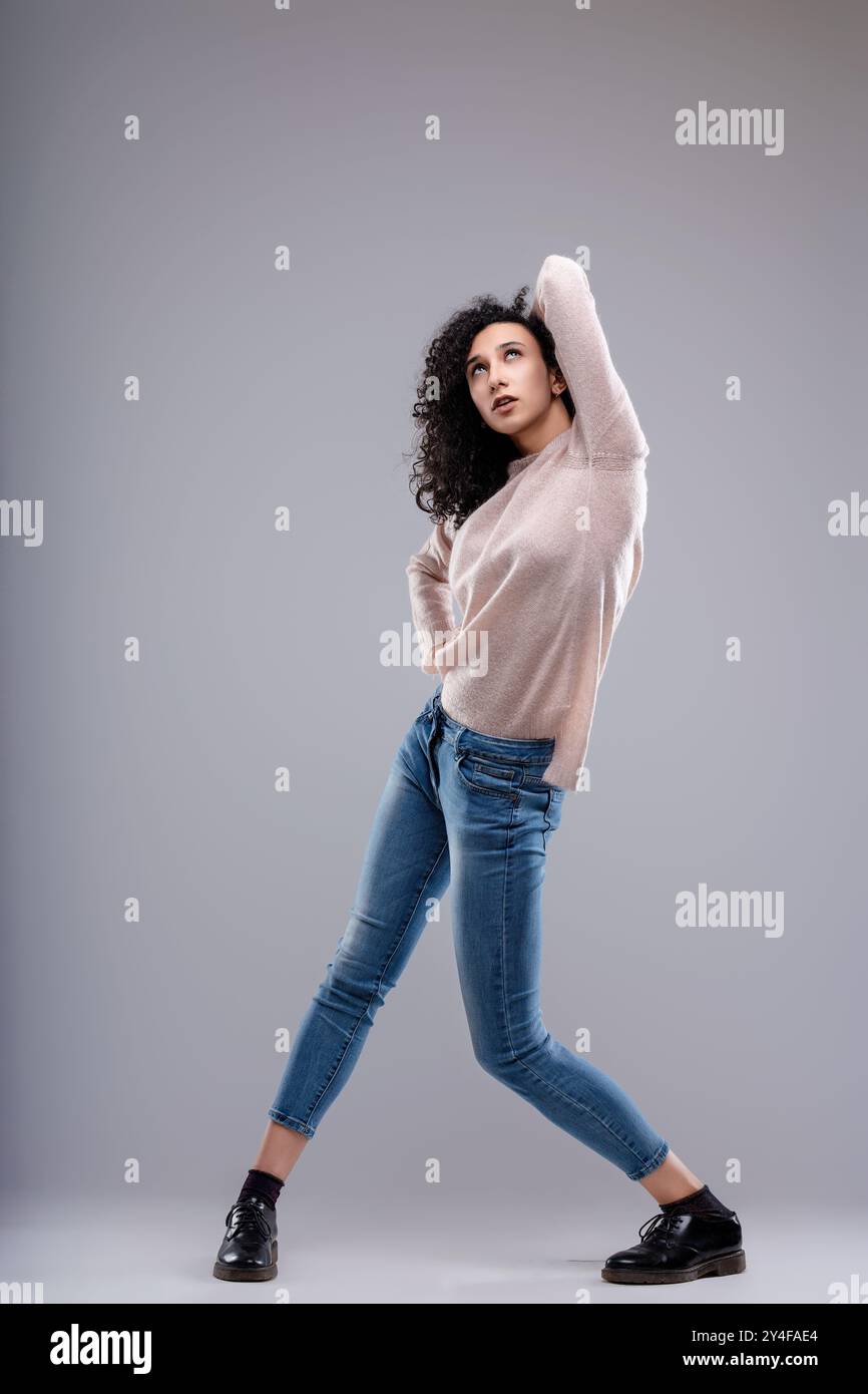 Jeune femme aux cheveux bouclés pose avec une main dans ses cheveux et l'autre sur sa hanche, ses jambes sont écartées et une jambe est pliée Banque D'Images