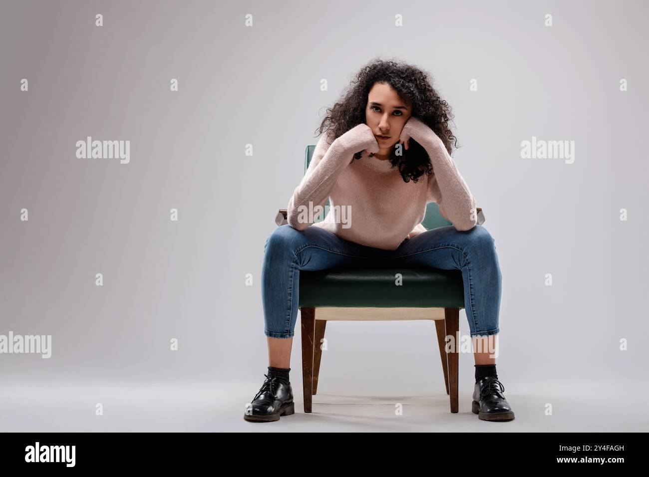 Jeune femme aux cheveux bouclés est assise sur une chaise avec ses coudes reposant sur ses genoux et son menton reposant sur ses mains, elle a l'air de s'ennuyer Banque D'Images