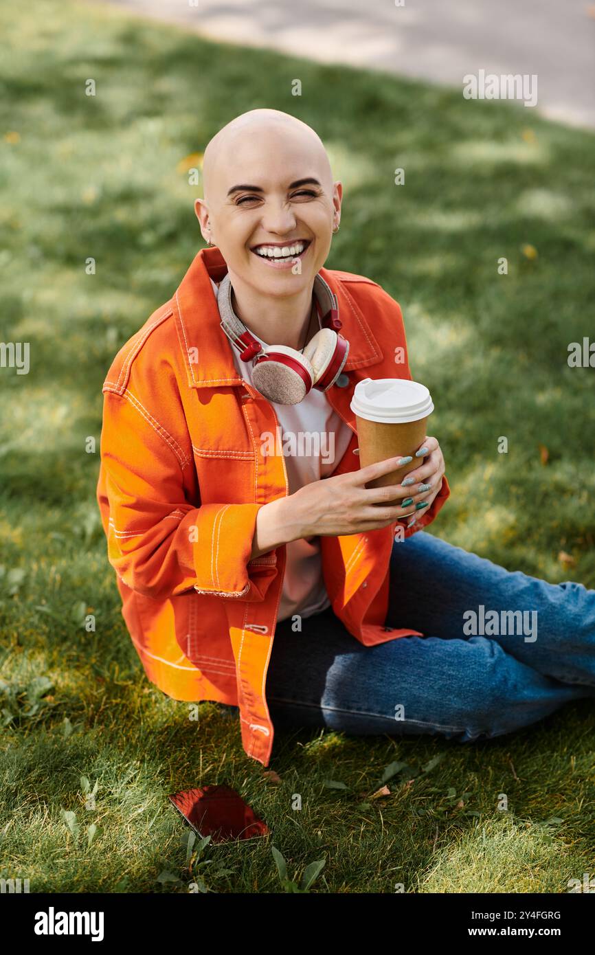 Une jeune femme chauve avec un sourire radieux sirote du café tout en étant assise sur l'herbe verte. Banque D'Images