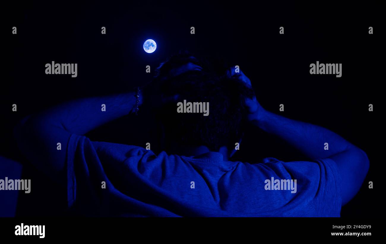 Une image saisissante capturée dans un moment de réflexion sous le clair de lune. Avec un homme sur le dos, tenant sa tête, et une teinte bleu profond, cette photo sym Banque D'Images