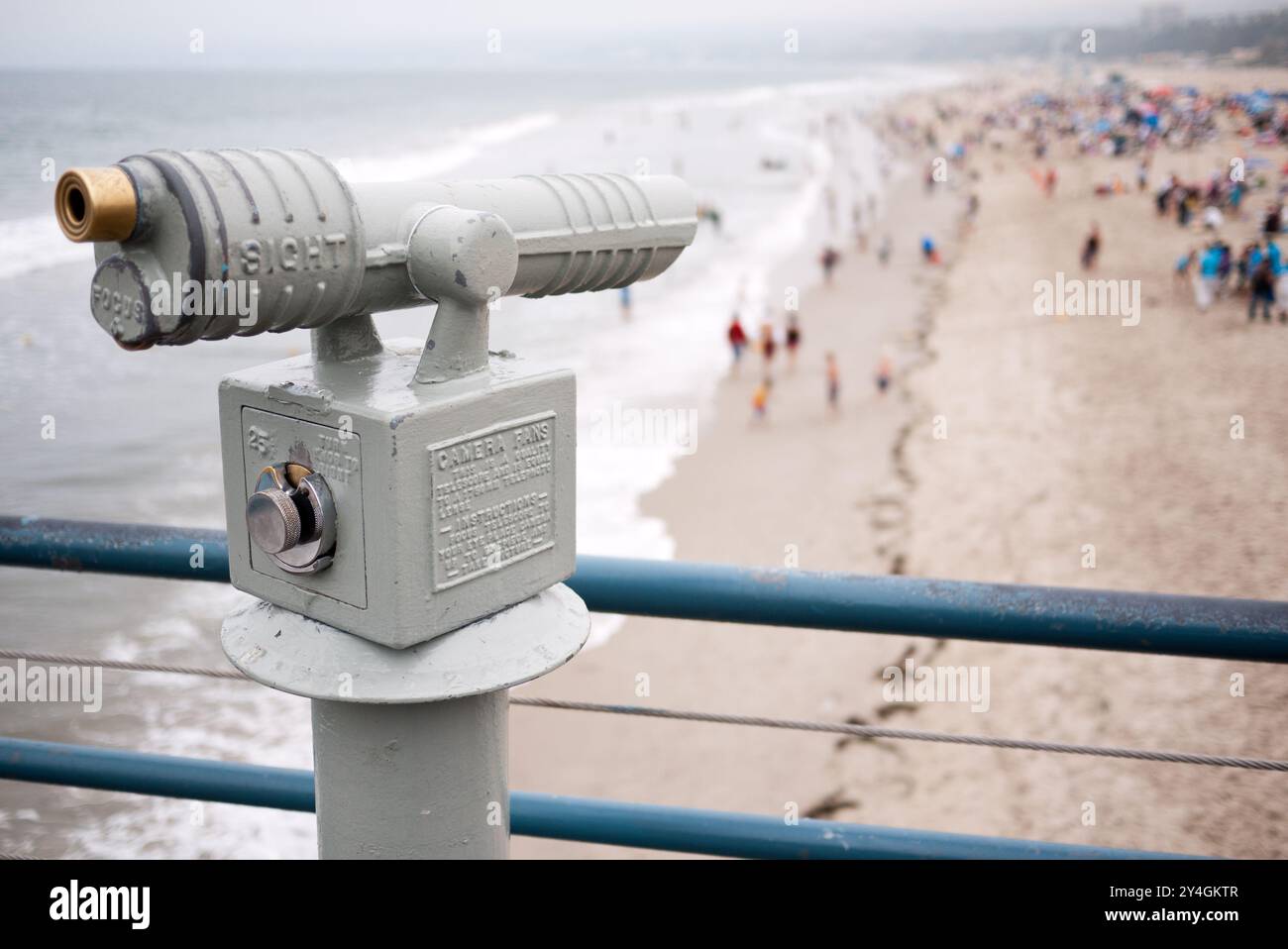 Santa Monica Pier coin Operated Telescope Santa Monica California // SANTA MONICA, États-Unis — Un télescope à pièces se trouve sur la jetée de Santa Monica, offrant aux visiteurs une vue sur l'océan Pacifique avec des foules de plage et des vagues visibles mais légèrement floues en arrière-plan. L'équipement de visualisation vintage est capturé avec une mise au point sélective, créant un bokeh atmosphérique qui suggère la scène vibrante de plage au-delà. La jetée de Santa Monica, construite en 1909, reste l'un des monuments les plus reconnaissables et des destinations touristiques populaires de la Californie du Sud. Banque D'Images