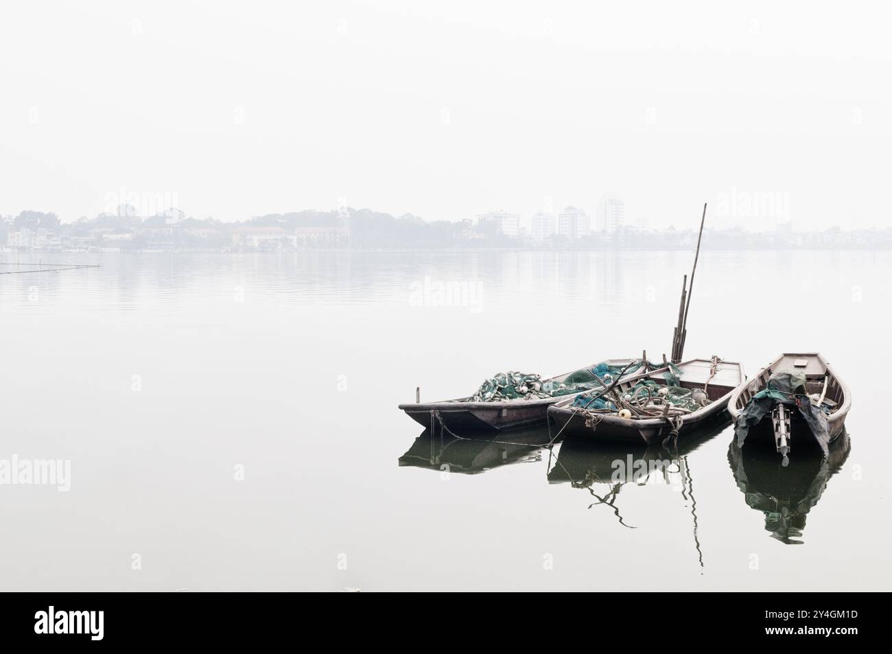 Sampans avec filets de pêche Lac de l'Ouest Hanoi Vietnam // HANOI, Vietnam — trois sampans en bois chargés de filets de pêche sont amarrés sur le Lac de l'Ouest (Ho Tay) à Hanoi, enveloppés par une brume épaisse. Les bateaux traditionnels contrastent avec le paysage urbain moderne, offrant un aperçu de la culture de pêche durable du Vietnam au milieu du développement urbain et des défis environnementaux. Banque D'Images