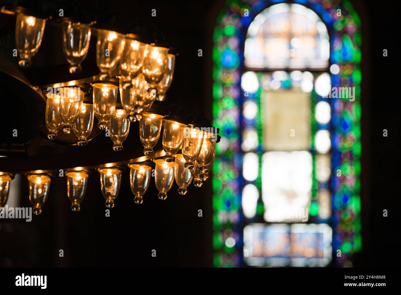 Hagia Sophia lustre ottoman vitrail Istanbul // ISTANBUL, Turquie — Un lustre ottoman traditionnel « top kandil » est suspendu devant un vitrail orné à Sainte-Sophie. Ces fenêtres en verre coloré, ajoutées lors des rénovations ottomanes, créent des motifs lumineux distinctifs dans l'espace sacré. La combinaison de sources lumineuses naturelles et artificielles représente une caractéristique de l'architecture des mosquées ottomanes. Banque D'Images