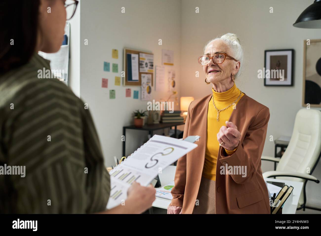 Femme d'affaires âgée conversant avec un collègue dans le bureau rempli de graphiques et d'affiches. Engager des discussions détaillées sur les stratégies et les développements commerciaux Banque D'Images