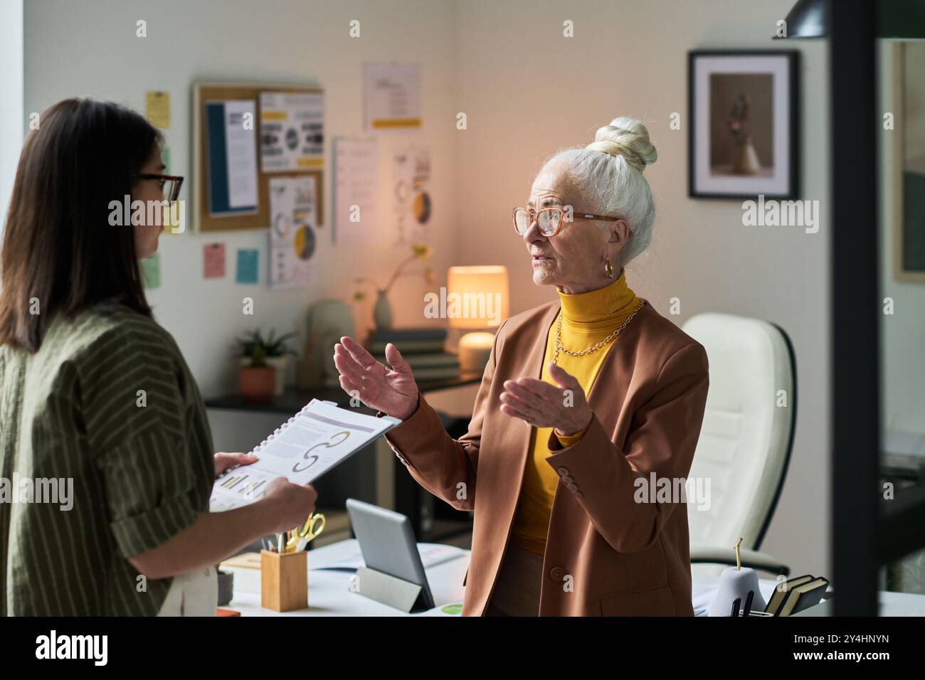 Femme d'affaires senior debout dans le bureau, engagé dans la discussion avec un collègue plus jeune tenant des documents, avec l'accent sur le travail d'équipe et la collaboration professionnelle, la création d'idées Banque D'Images