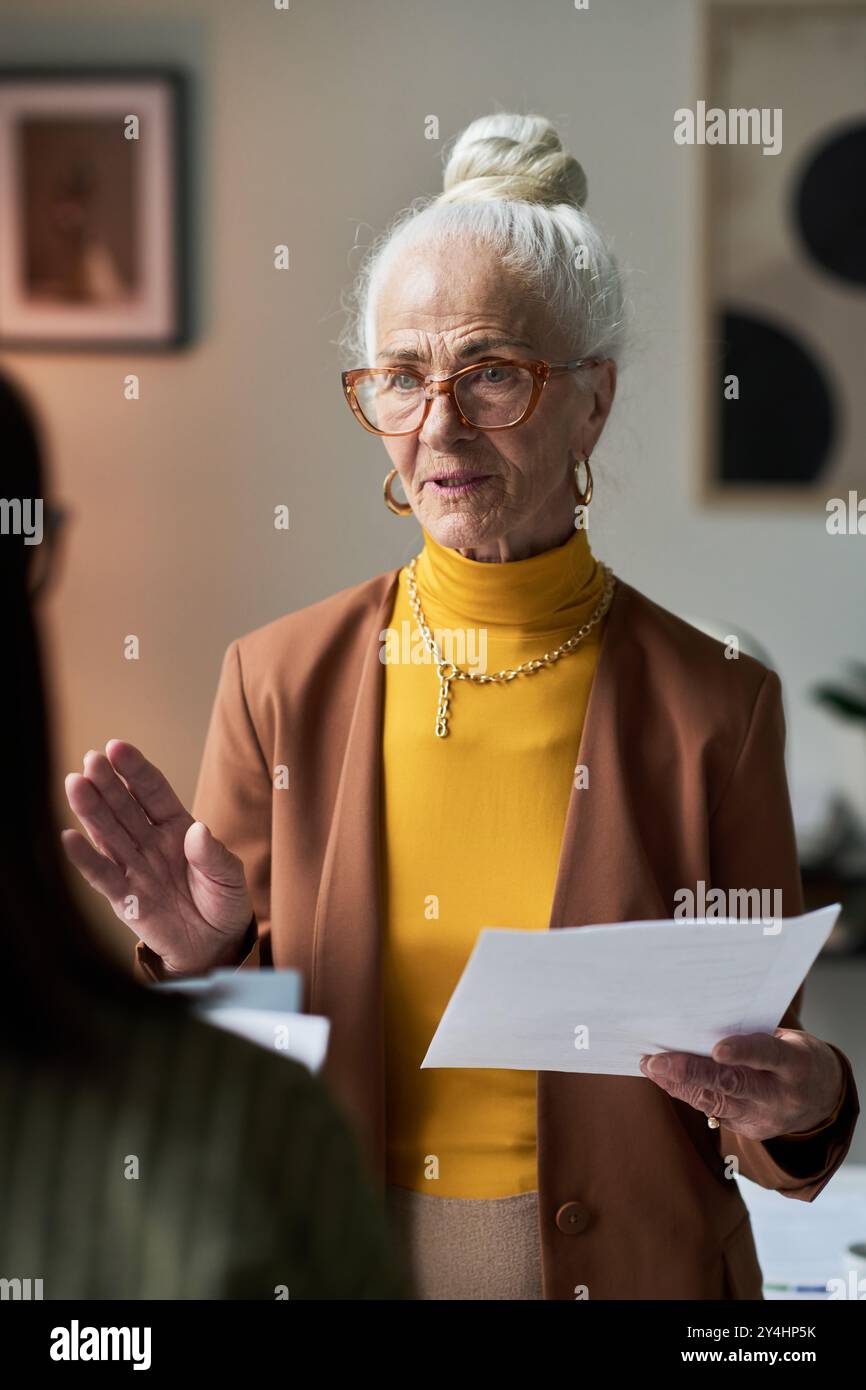 Femme âgée avec des lunettes tenant des papiers blancs tout en étant engagée dans la discussion dans un cadre de bureau contemporain avec des pièces d'art encadrées et des meubles modernes en arrière-plan Banque D'Images