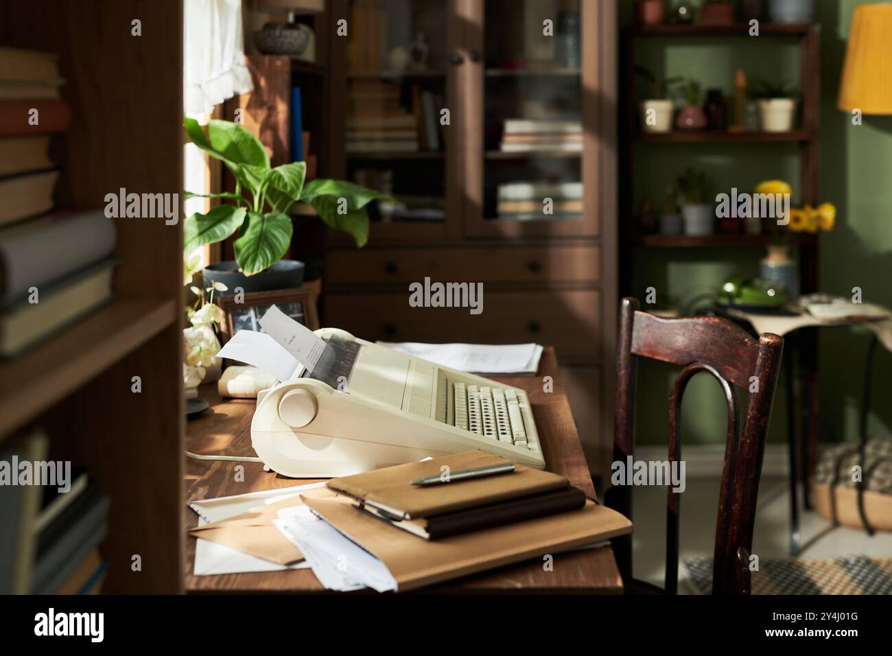 Configuration de bureau à domicile vintage avec une vieille machine à dactylographier, bureau en bois, diverses plantes d'intérieur, livres et cahiers. Lumière du soleil éclairant la pièce à travers des stores partiellement ouverts Banque D'Images