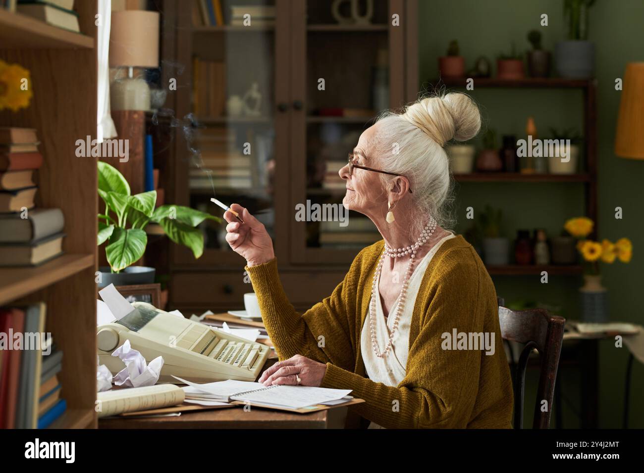 Femme âgée avec les cheveux blancs attachés, tenant une cigarette, travaillant dans un environnement de bureau avec des livres, des plantes et la technologie rétro qui l'entourent Banque D'Images