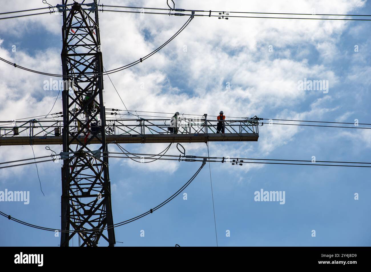Travailleurs de lignes électriques à haute tension effectuant des travaux de maintenance sur une tour de transmission contre un ciel bleu avec des nuages dispersés Banque D'Images