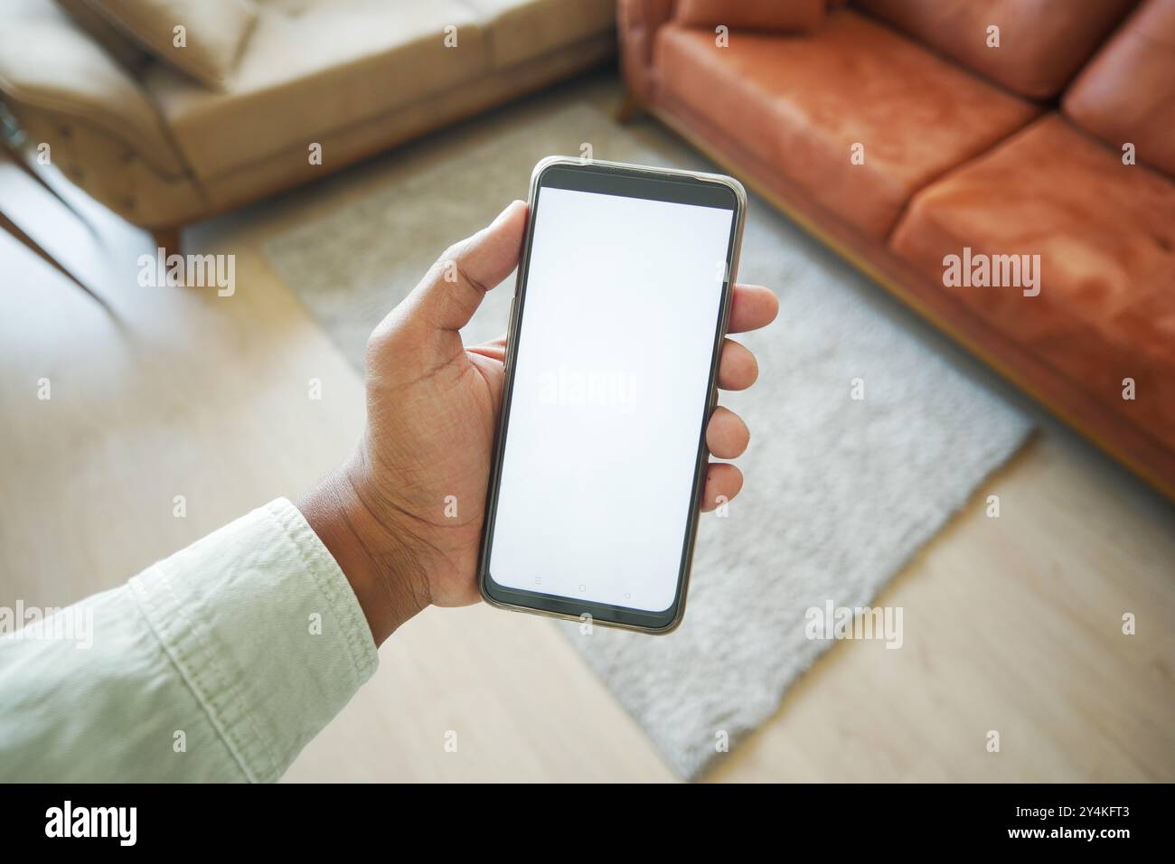 Une main tenant fermement un smartphone doté d'un écran vierge dans un espace salon confortable Banque D'Images
