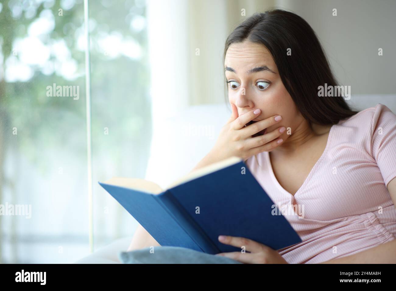 Femme surprise lisant un livre de papier assis sur un canapé dans une maison Banque D'Images