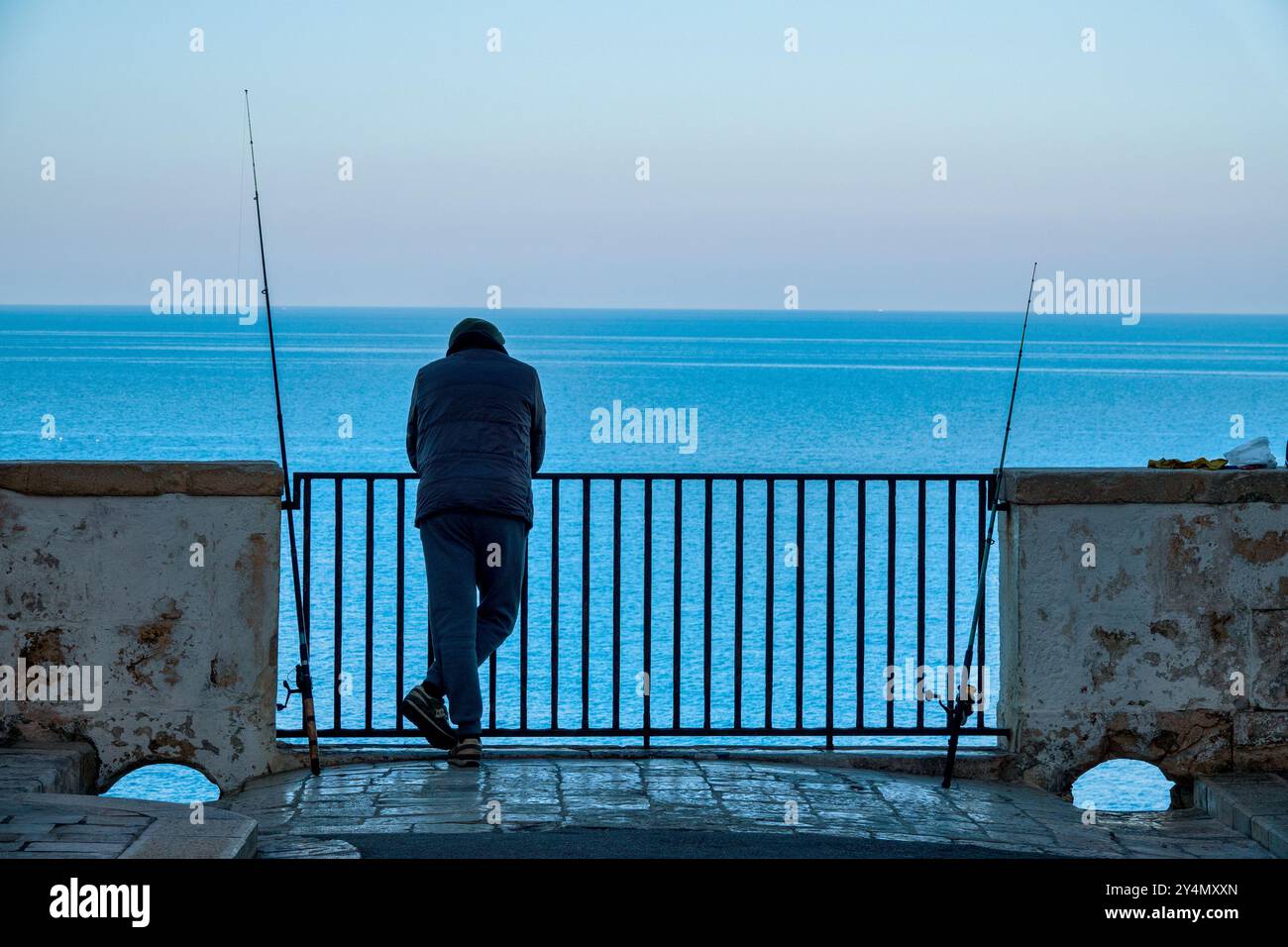 Un pêcheur debout près de la rampe à Polignano a Mare, Italie, avec des cannes à pêche à ses côtés. Banque D'Images