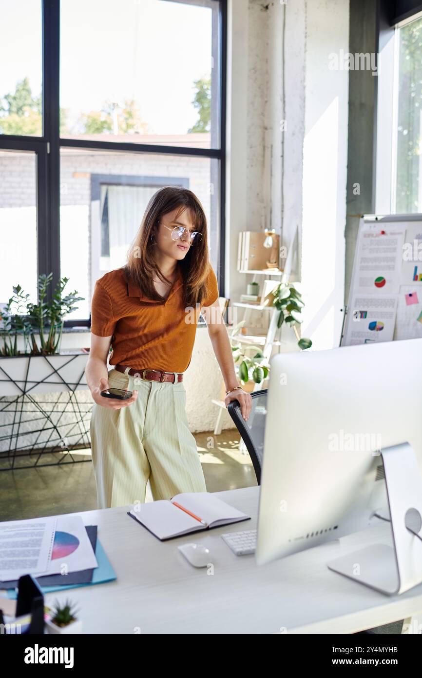 Une jeune personne non binaire examine son ordinateur tout en se tenant debout dans un bureau lumineux. Banque D'Images