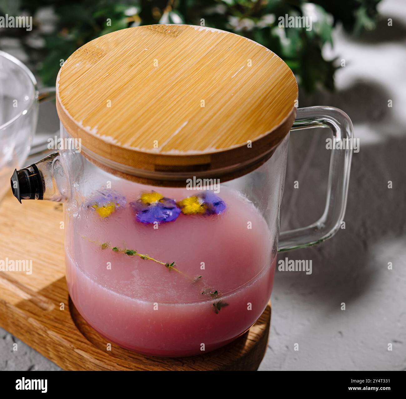 Théière en verre transparent avec tisane rose et fleurs comestibles sur un plateau en bambou, stylisée sur un fond de béton Banque D'Images