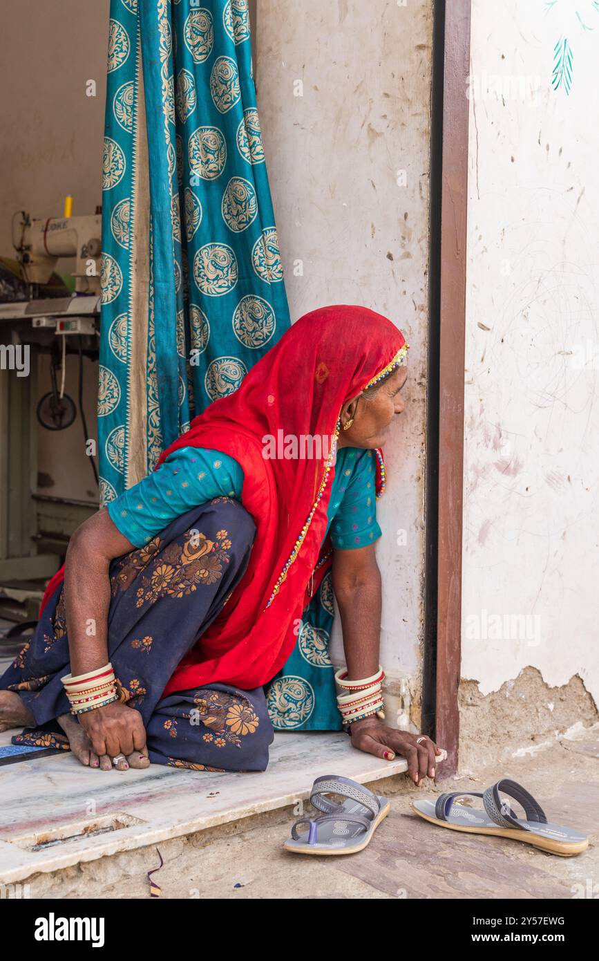 Tilora, Ajmer, Rajasthan, Inde. 9 novembre 2022. Femme dans un foulard rouge regardant hors d'une porte. Banque D'Images