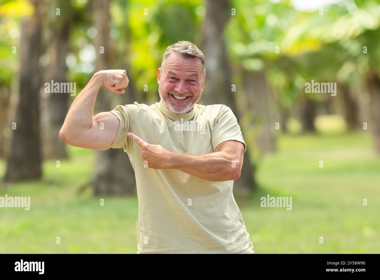 Un homme mûr joyeux fléchissant le bras et pointant vers ses biceps tout en souriant brillamment, profitant d'un moment amusant en plein air dans un cadre verdoyant et ensoleillé. Banque D'Images