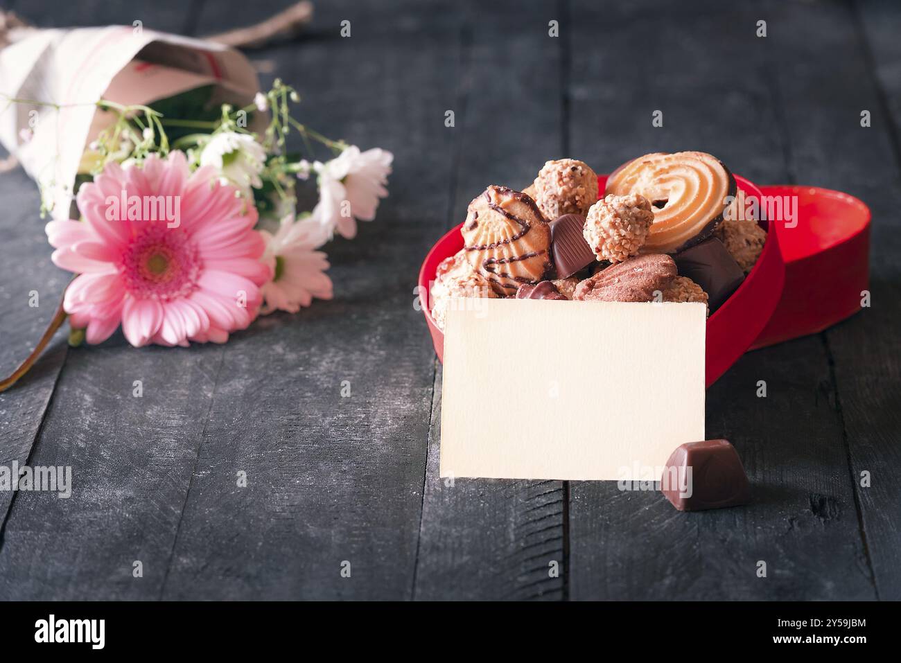 Idée de carte de félicitations avec une note de papier vide appuyée contre une boîte rouge pleine de biscuits, chocolat et un bouquet de fleurs en arrière-plan, sur Banque D'Images