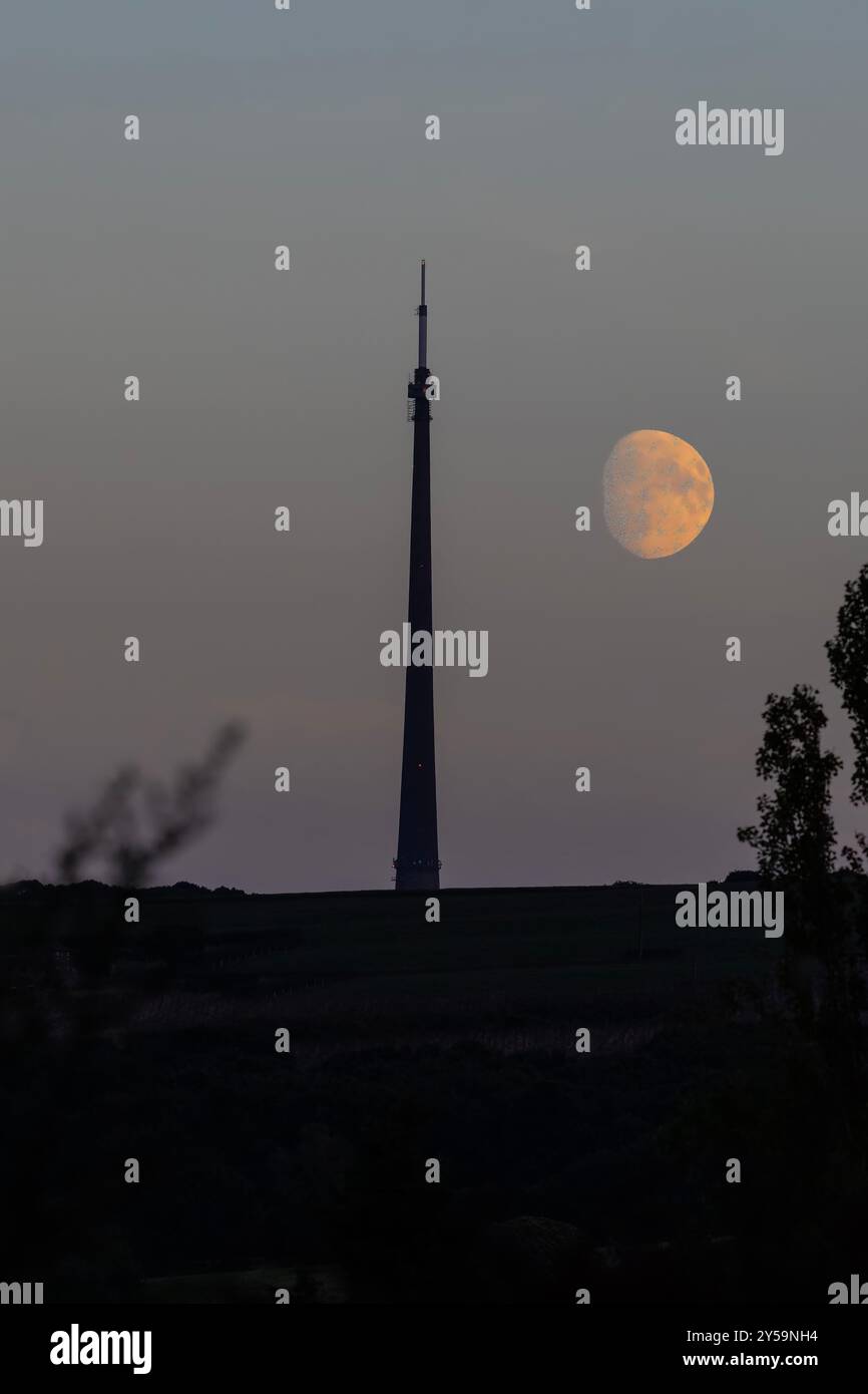 Une grande pleine lune devient visible au crépuscule derrière le contour sombre de la tour Arqiva ou du mât de transmission Emley Moor près du village Emley. Banque D'Images