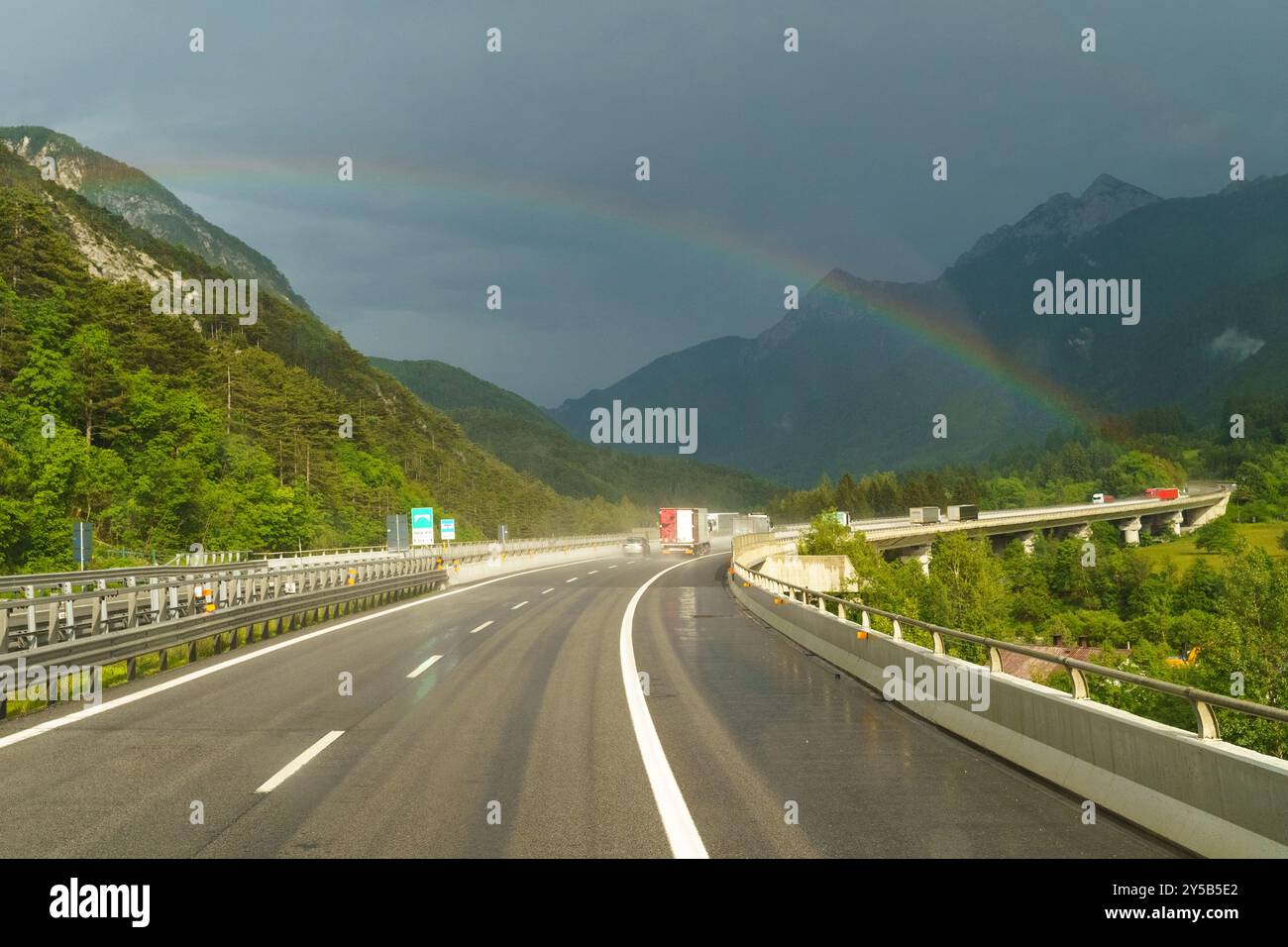 Une autoroute sinueuse traverse des montagnes verdoyantes, où les gouttes de pluie brillent sous la lumière pâle du soleil. Un arc-en-ciel éclatant arque magnifiquement, ajoutant de la couleur Banque D'Images