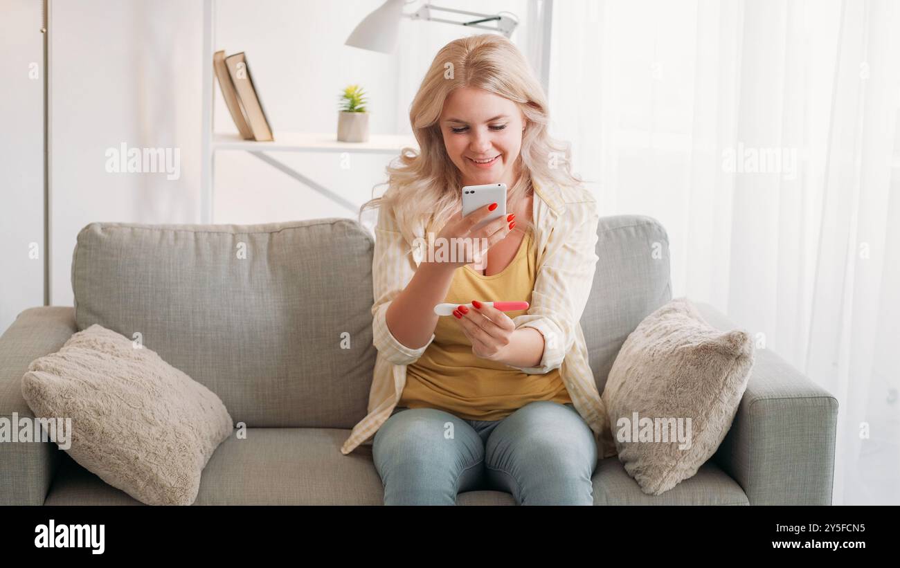Grossesse désirable. Femme heureuse. Partage des actualités. Inspirée dame souriante prenant une photo du résultat positif du test sur l'intérieur de la pièce lumineuse du canapé assis du smartphone. Banque D'Images