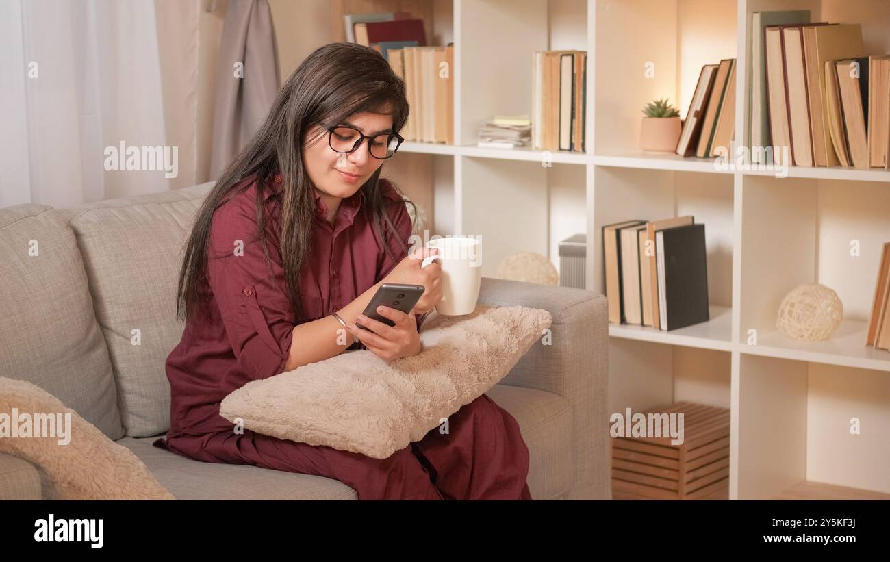 Café détendez-vous. Divertissement sur smartphone. Loisirs à domicile. Heureuse femme rêveuse buvant tasse de thé discutant sur cellule profitant du repos de week-end confortable sur le canapé dans le salon. Banque D'Images
