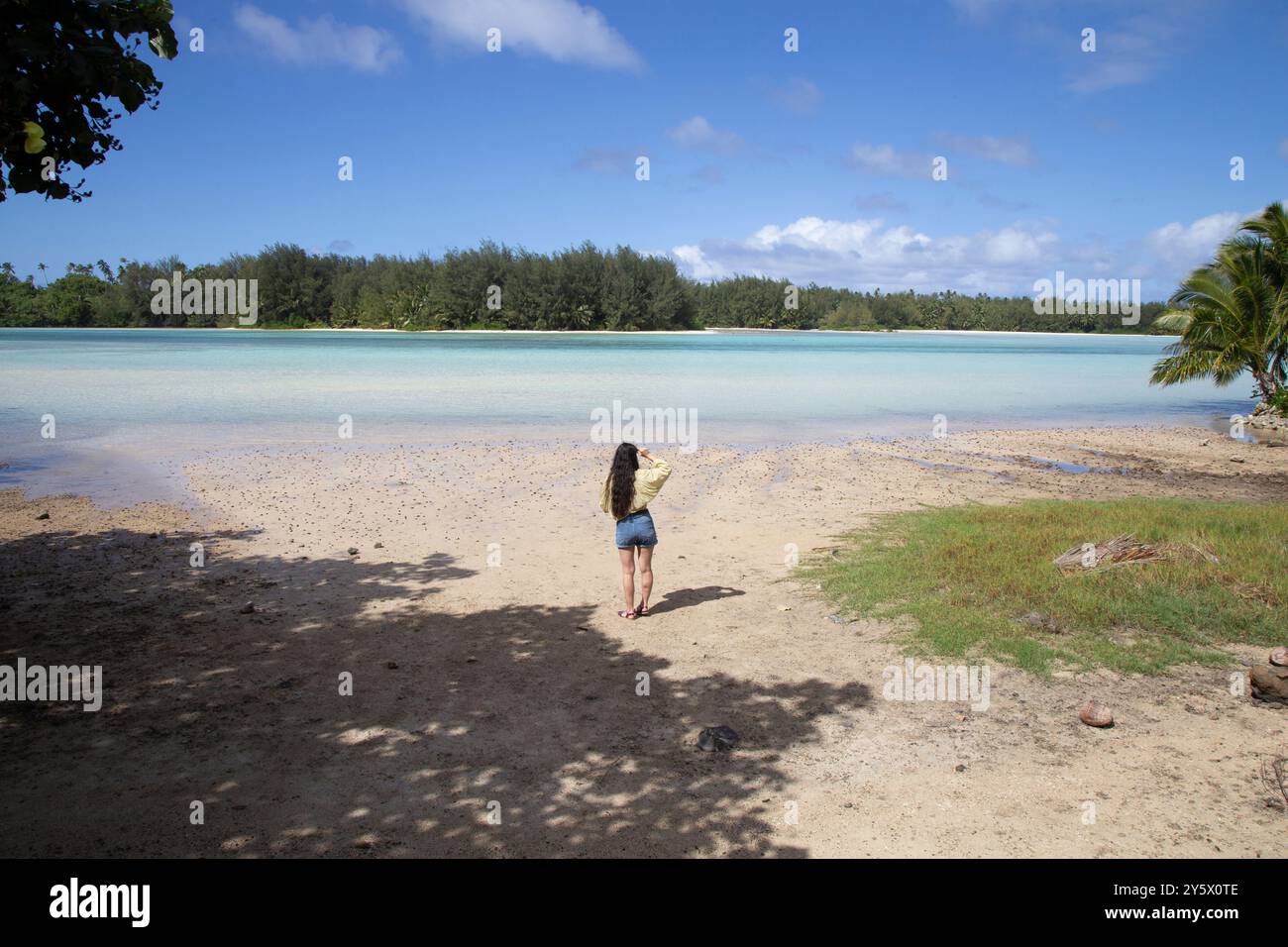 Une scène de plage tropicale sereine avec une femme debout sur le sable regardant sur les eaux bleues calmes et le feuillage vert au loin, Muri, Rarotonga, Îles Cook Banque D'Images