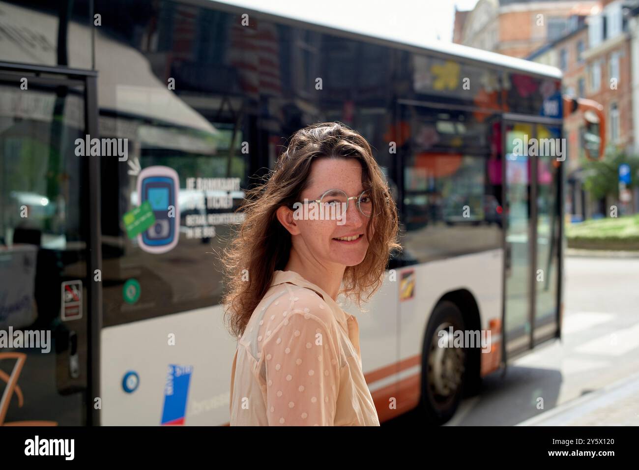 Femme souriante dans des lunettes debout devant un bus de la ville par une journée ensoleillée. Banque D'Images