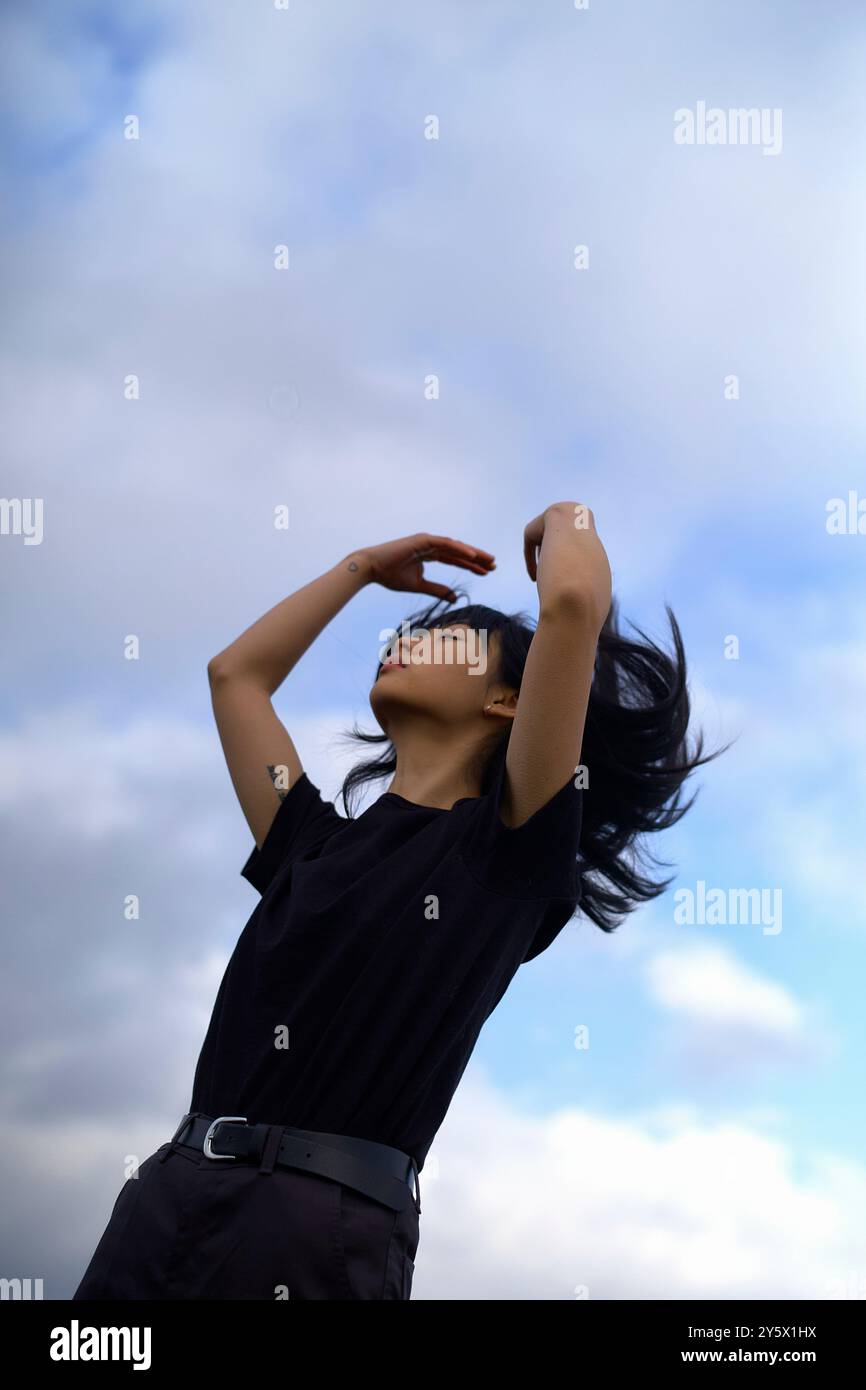 Une femme aux cheveux noirs regarde vers le haut contre un fond de ciel nuageux, sa main touchant gracieusement ses cheveux. Banque D'Images