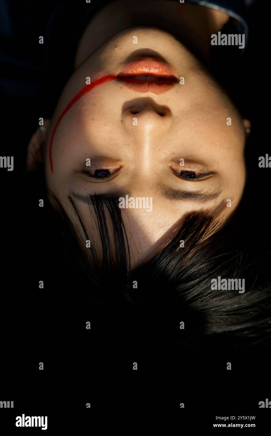 Portrait en gros plan d'une femme à l'envers avec une strie rouge sur le front et un maquillage saisissant dans un éclairage dramatique. Banque D'Images
