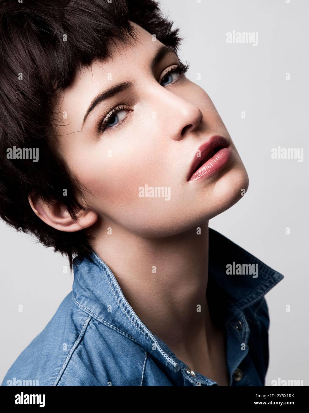 Portrait en gros plan d'une femme aux cheveux bruns courts et au maquillage, portant une chemise bleue, regardant loin sur un fond clair. Banque D'Images