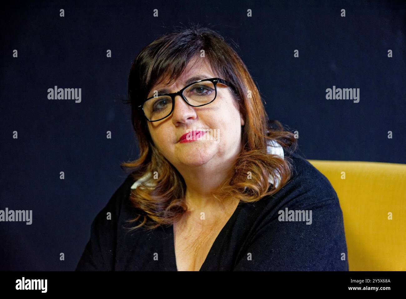 Femme d'âge moyen confiante avec des lunettes et des cheveux aux épaules posant sur un fond noir dans une chaise jaune. Banque D'Images