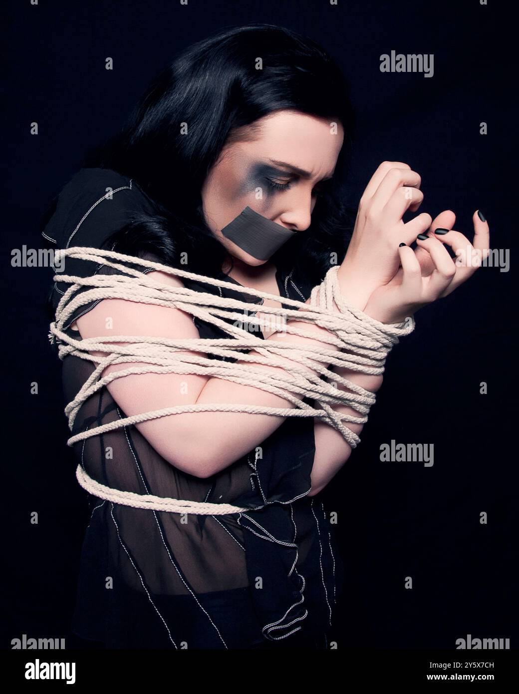 Femme avec les cheveux foncés attaché avec une corde sur un fond noir, regardant ses mains liées. Banque D'Images