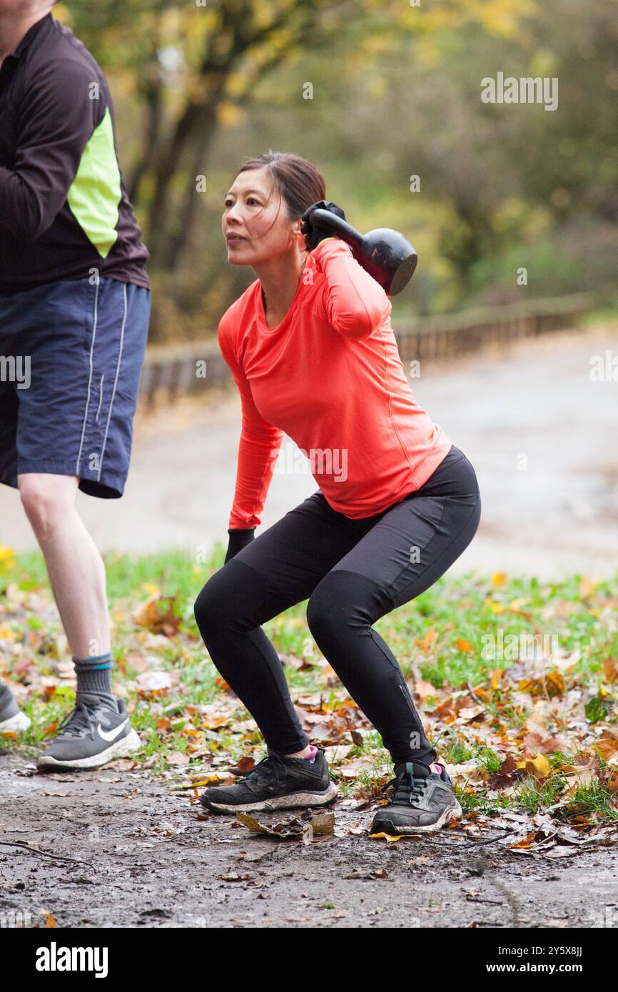 Femme en haut orange effectuant des exercices de kettlebell dans un parc extérieur avec des feuilles d'automne sur le sol et une vue partielle d'un homme en tenue de sport. Banque D'Images