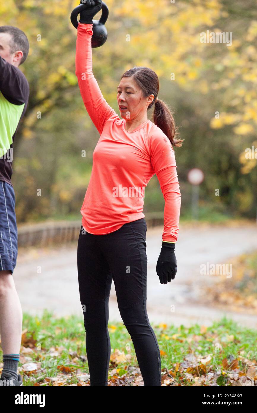 Une femme fait de l'exercice avec des kettlebells dans un parc pendant l'automne tandis qu'une autre personne s'entraîne à proximité. Banque D'Images
