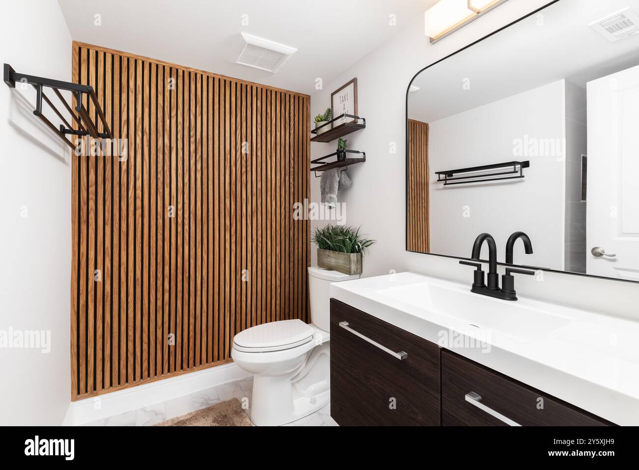 Une salle de bain avec une armoire en bois sombre, des étagères flottantes avec des décorations au-dessus des toilettes et un mur d'accent à lattes en bois. Banque D'Images