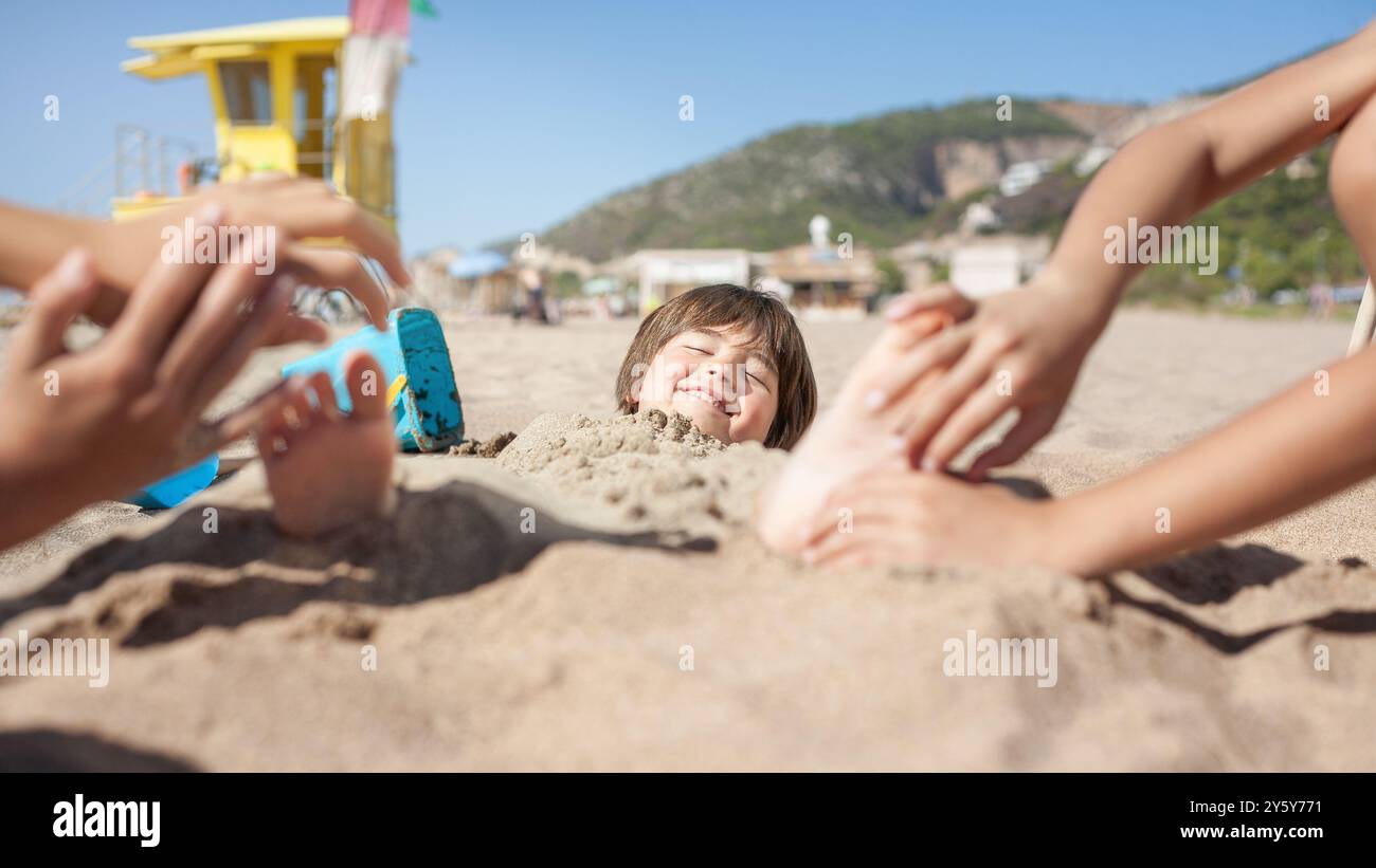 Garçon couvert de sable sourit tandis que les mains chatouillent la plante de ses pieds. Banque D'Images