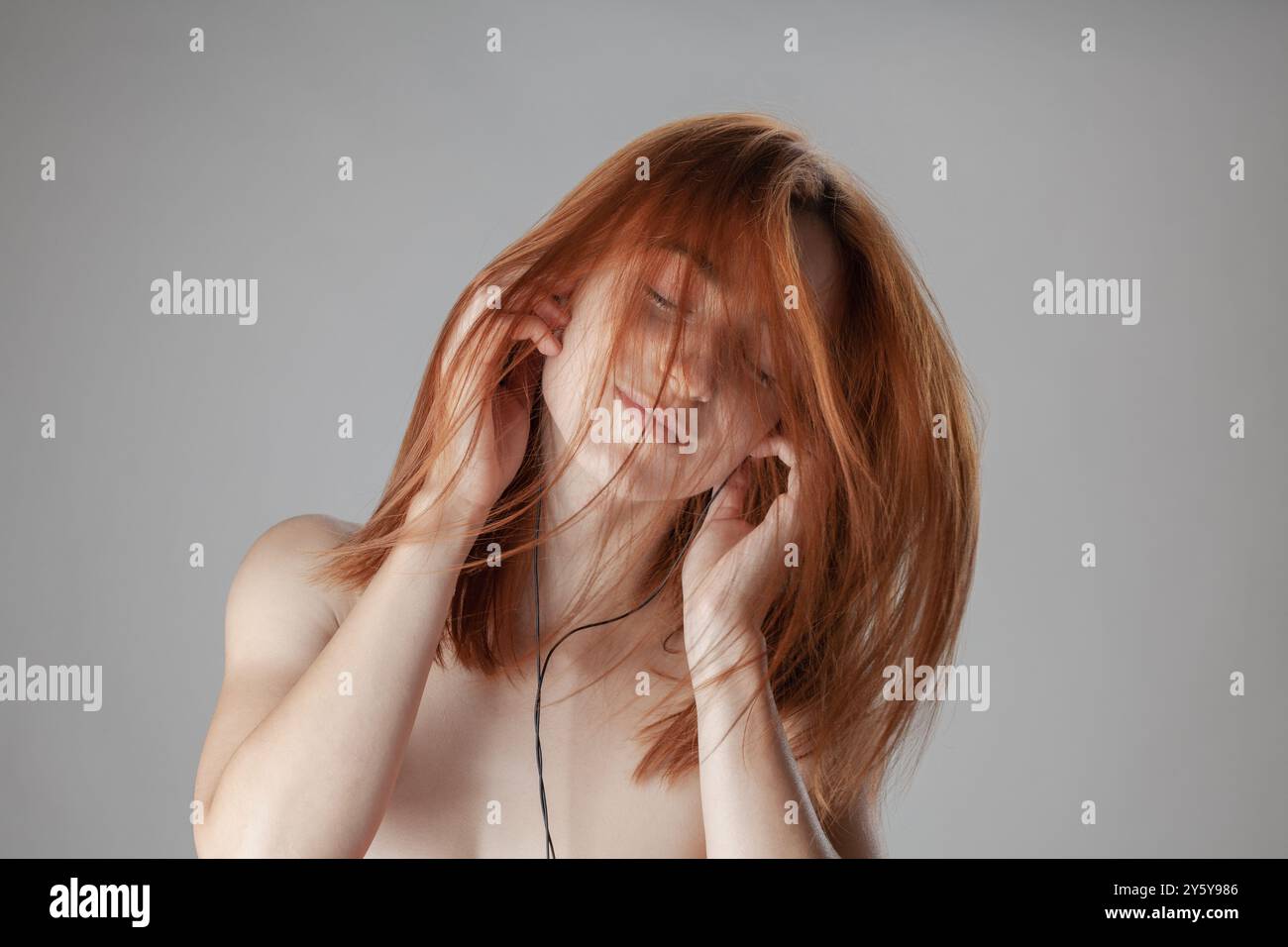 Femme rousse dans un moment serein, les yeux fermés, touchant ses cheveux doucement sur un fond gris, véhiculant des sentiments de paix et de connexion de soi. Banque D'Images