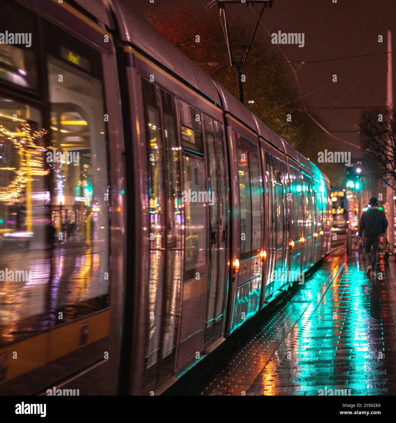 Photo de nuit de Dublin avec un tramway Luas par une soirée pluvieuse, encadrée à travers les ordures, reflétant les lumières de la ville sur les rues humides avec des gouttes de pluie. Banque D'Images